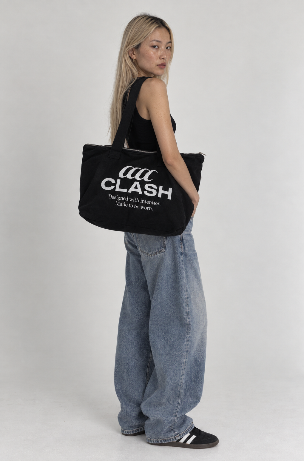 A woman stands in profile holding a large black tote bag with bold white 'CLASH' branding and smaller text. She is wearing a black sleeveless top, loose light-wash blue jeans, and black sneakers with white stripes, against a plain light gray studio background.