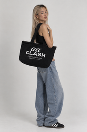A woman modeling a black canvas tote bag with white 'CLASH' branding, styled with light-wash wide-leg jeans and black sneakers against a plain light gray studio background.