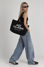 A woman wearing a black tank top, wide-leg light-wash jeans, black sneakers, and black sunglasses carries a black canvas tote bag with white 'CLASH' branding printed on the side, set against a plain light gray studio background.
