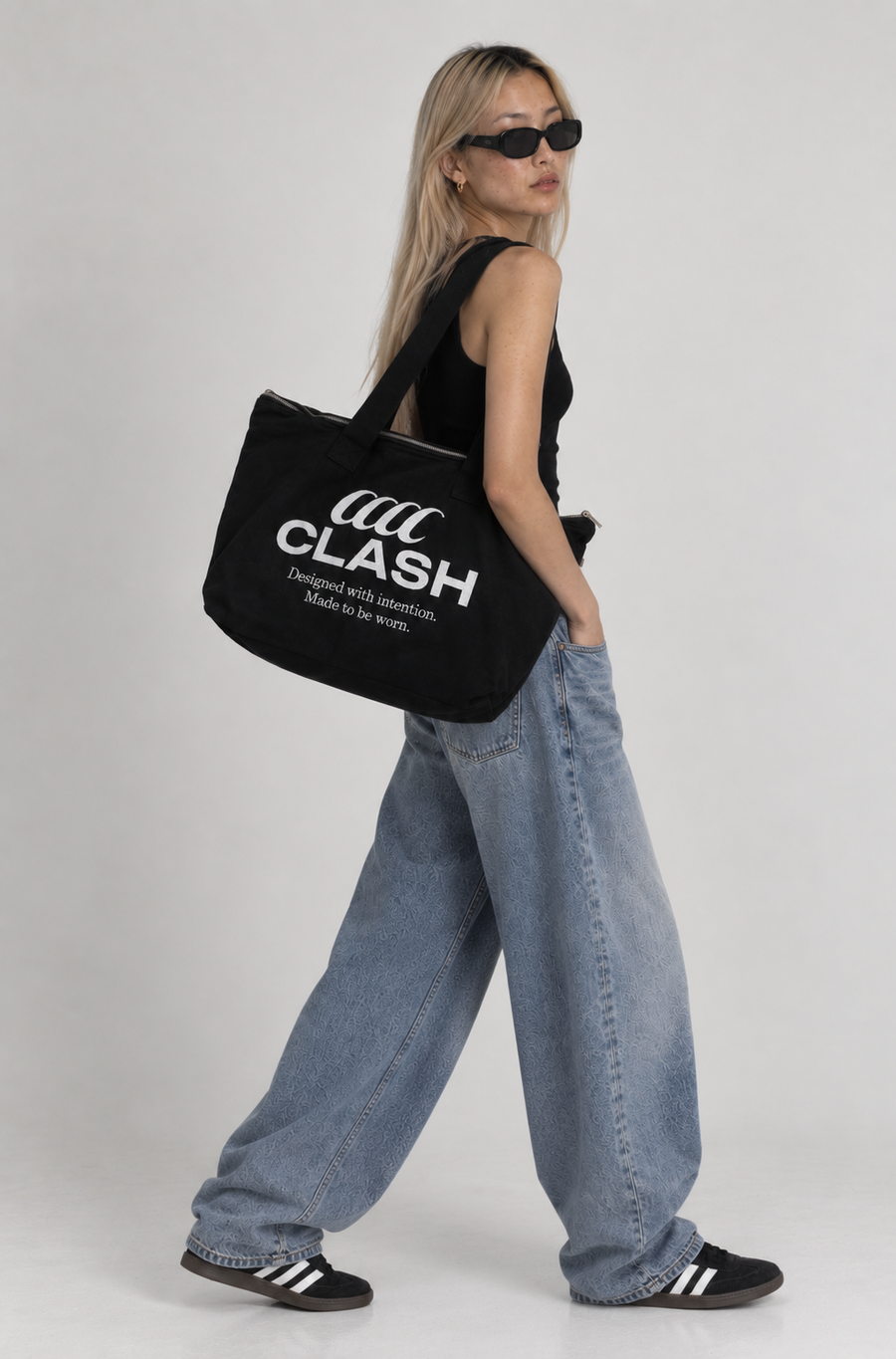 A woman wearing a black tank top, wide-leg light-wash jeans, black sneakers, and black sunglasses carries a black canvas tote bag with white 'CLASH' branding printed on the side, set against a plain light gray studio background.