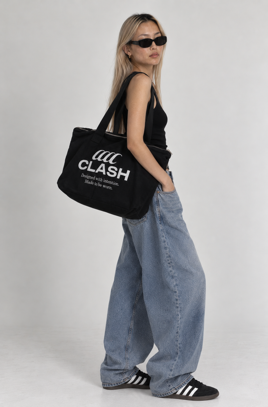 A woman wearing a black tank top, wide-leg light blue jeans, black sneakers, and sunglasses carries a large black tote bag with white text that reads 'CLASH' and smaller text below. She is photographed against a plain light gray studio background.