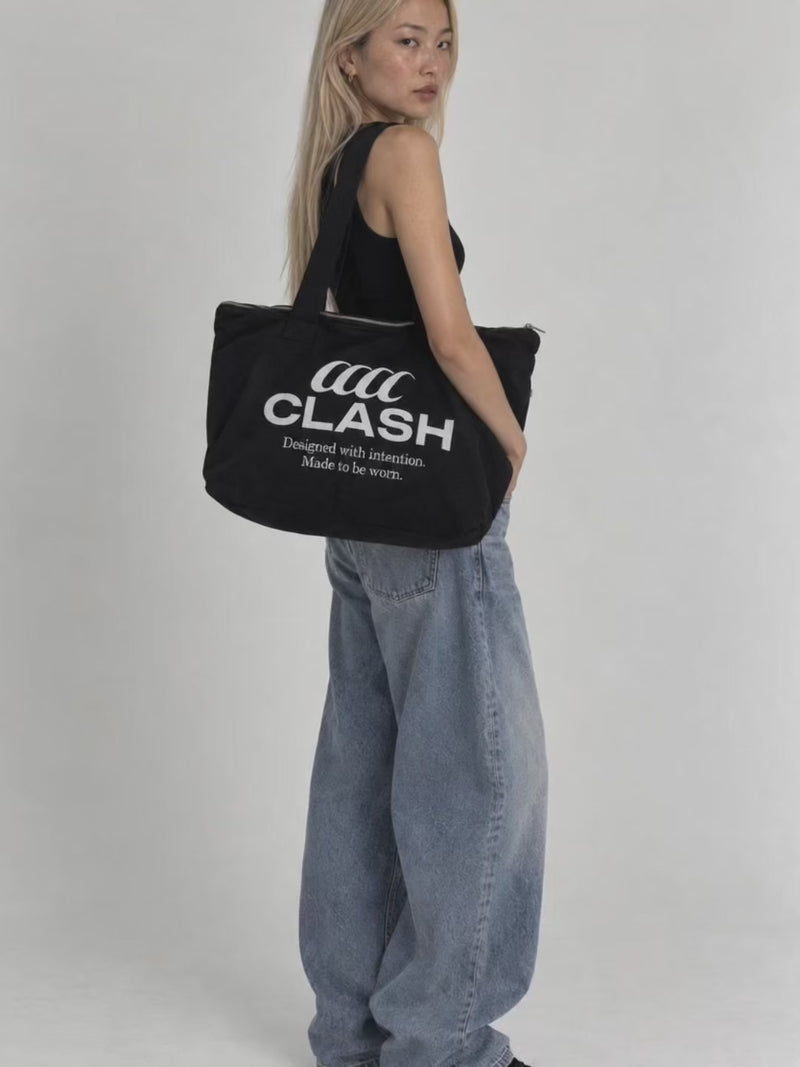 Model wearing the black Clash Foundation Tote Bag with embroidered logo, black tank top and baggy jeans in a minimalist studio.